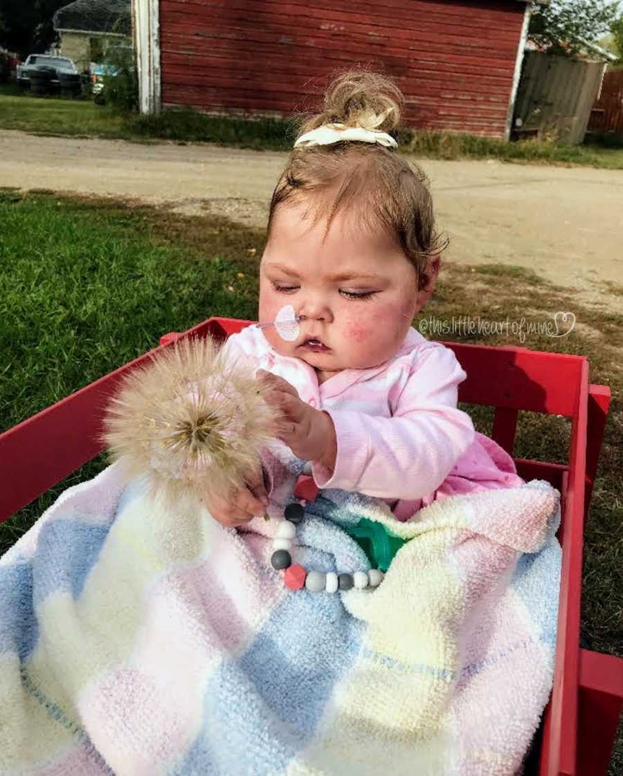 Little girl with tube up her nose sits in red wagon outside holding onto dandelion