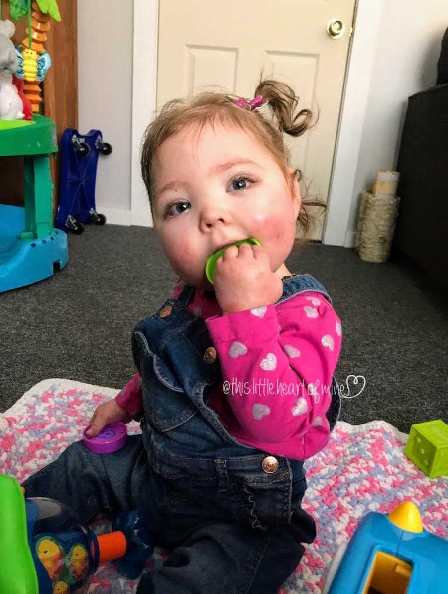 Baby sits on floor of home playing with toys holding toy to her mouth