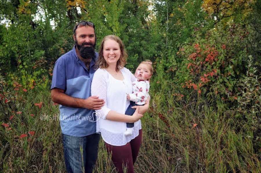 Husband stands beside wife in field who holds their daughter with heart issues