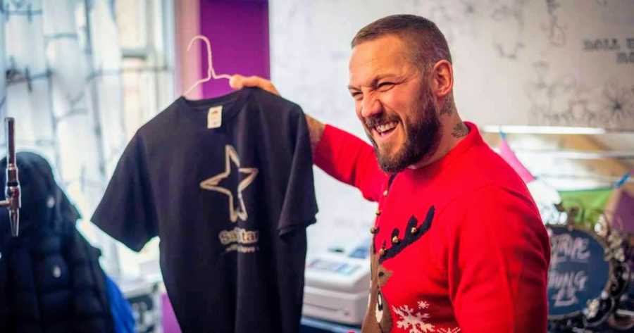 Man who is a recovered addict stands smiling in Christmas sweater as he holds up t-shirt on hanger