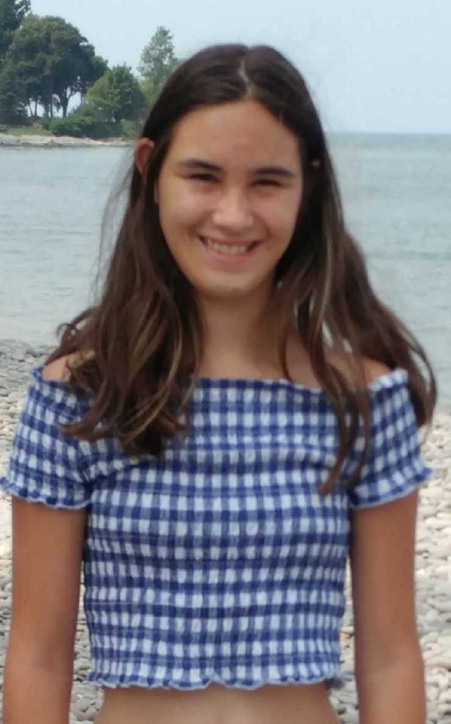 Young girl with two mothers stands smiling on beach near body of water