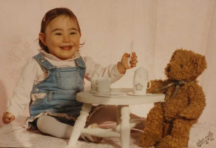 Baby girl smile as she sits at small table with tea set beside teddy bear