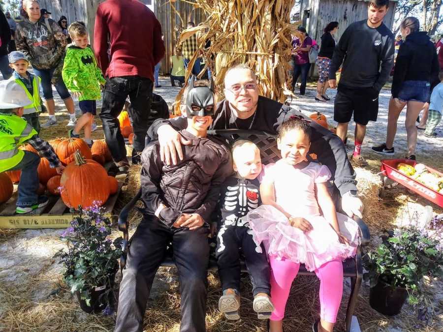 Father crouches behind bench where his three children sit in Halloween costumes
