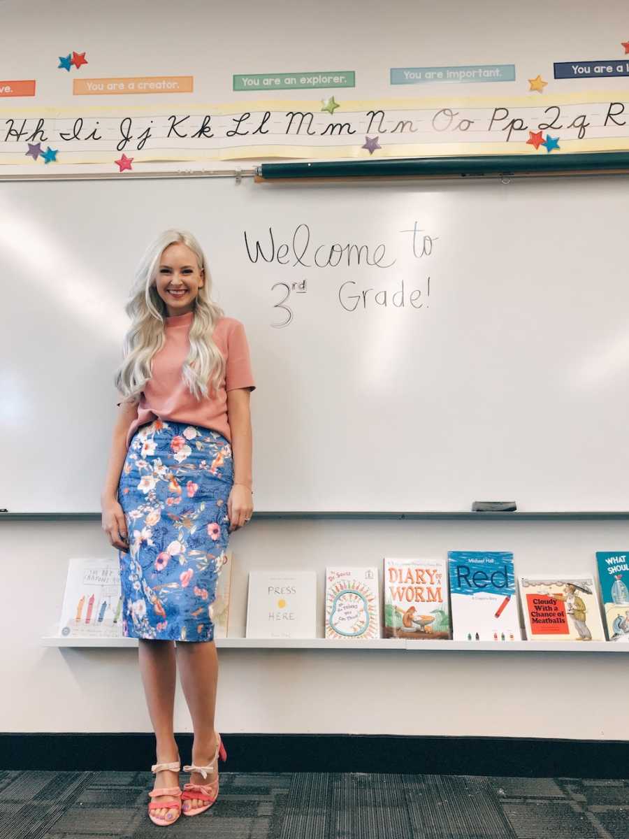 Woman stands in classroom beside white board that says, "Welcome to 3rd Grade"