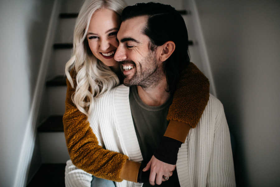 Husband sits on stairs smiling as wife wraps her arms around him
