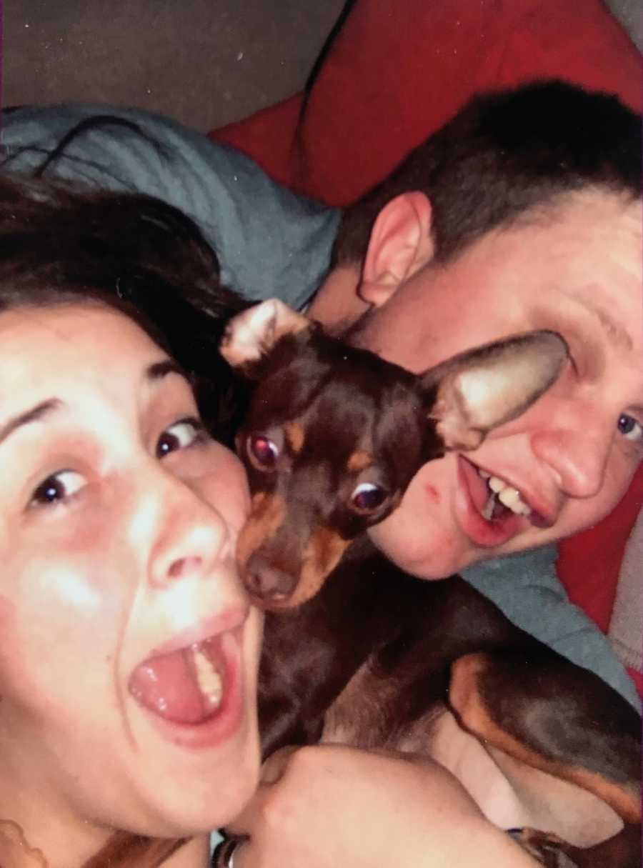 Husband and wife smile in selfie as their little dog lays between them