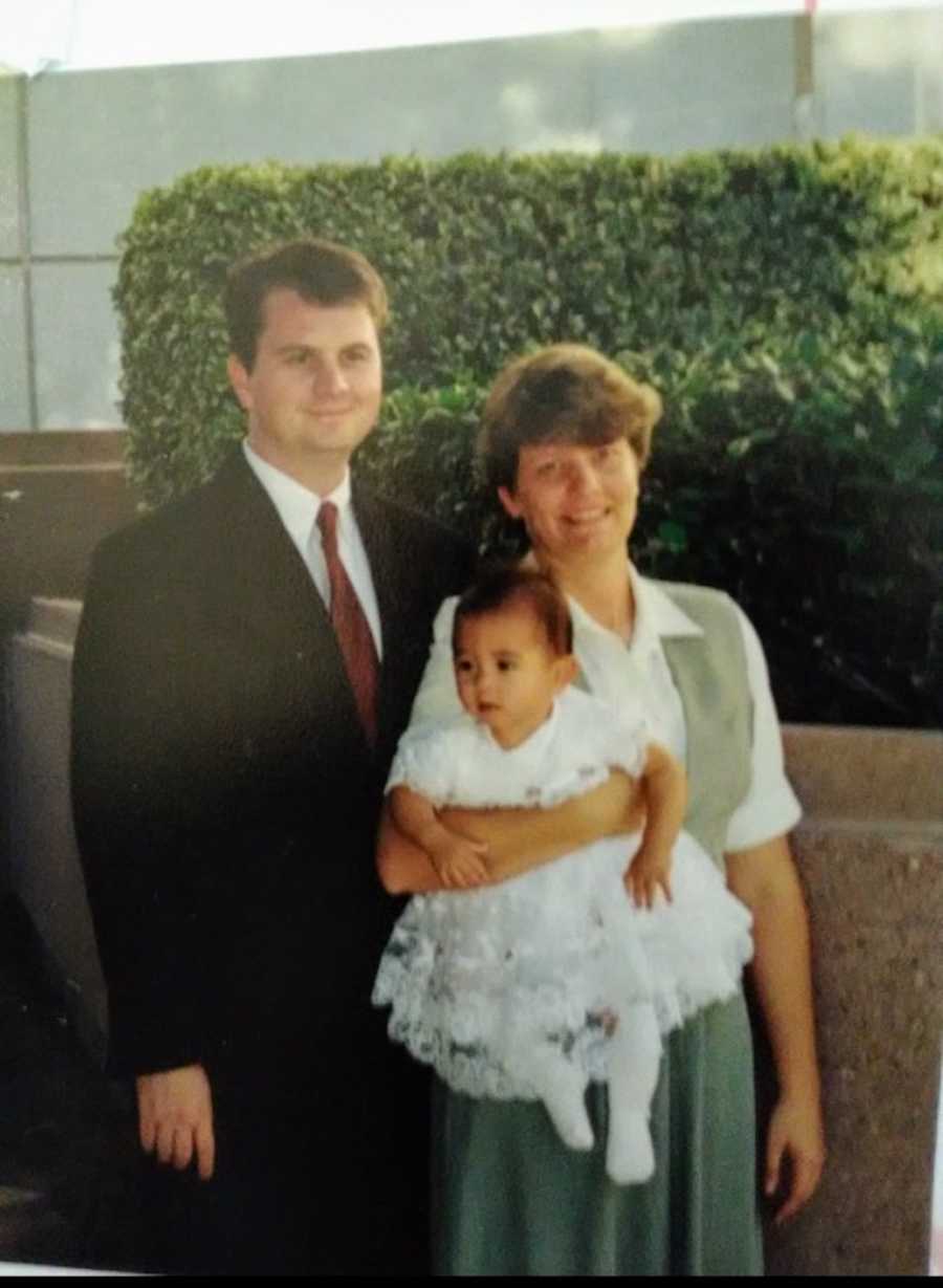 Mother smiles outside as she holds her adopted daughter beside her husband