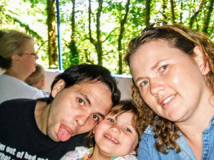 Woman takes selfie outside with her smiling daughter and husband who sticks his tongue out