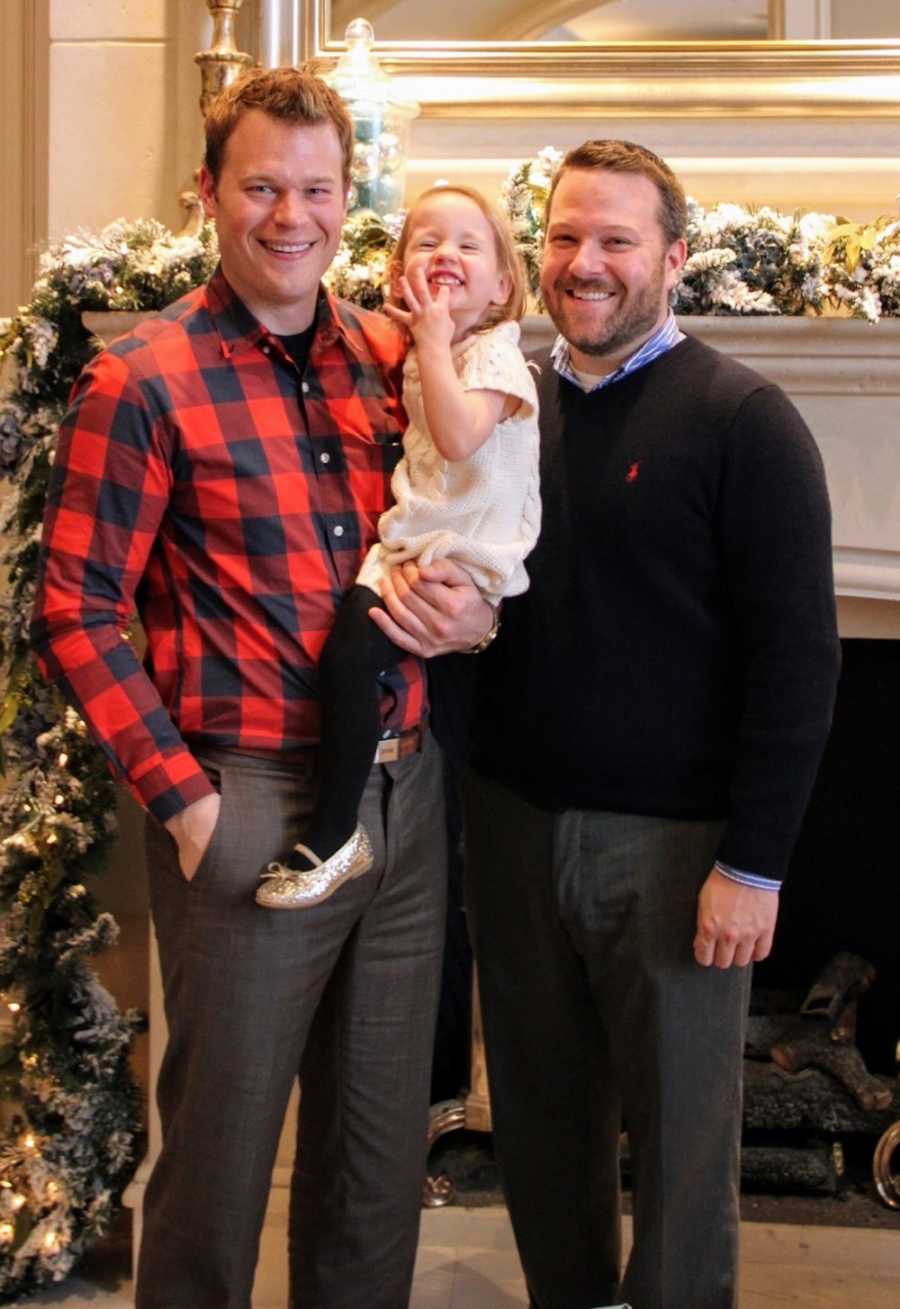 Husbands stand smiling in front of chimney decorated for Christmas with their daughter