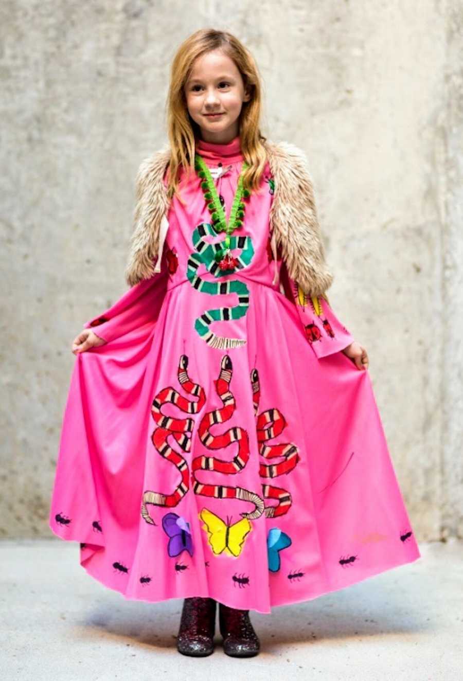 Little girl with two dads stand in pink dress with snakes on it with fur vest over