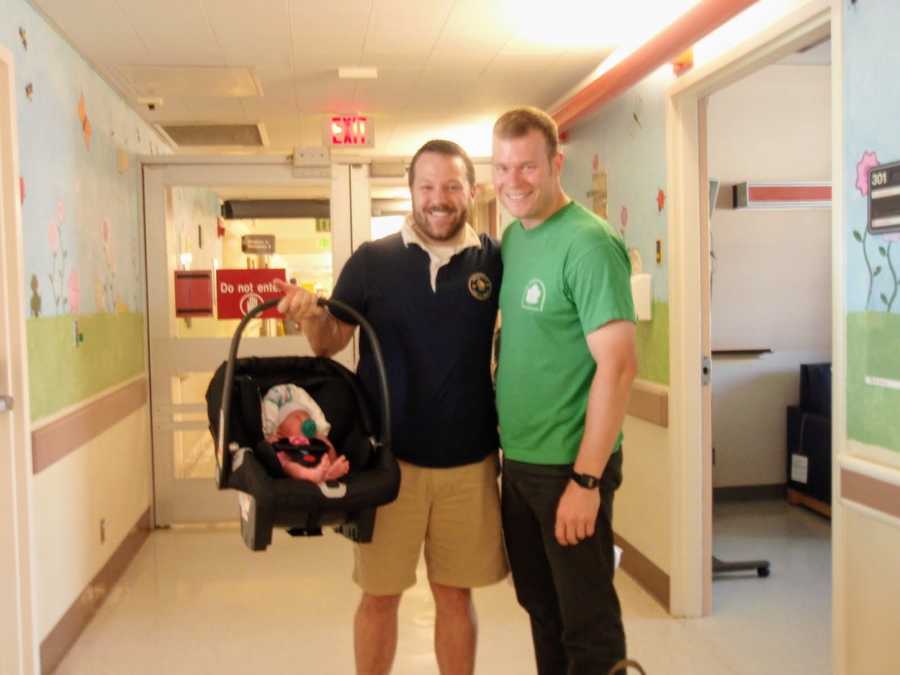Husbands stand smiling in hospital hallway as one holds carseat with adopted daughter