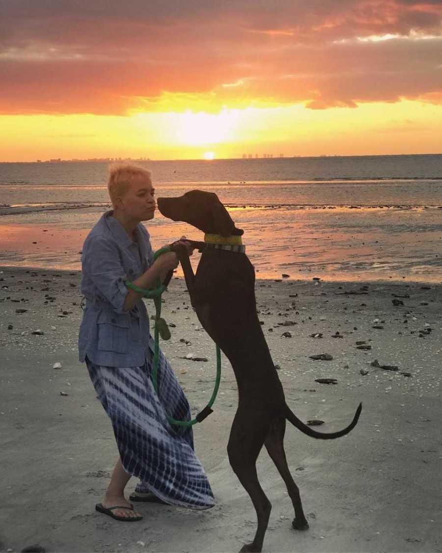 Woman who is back on her medication stands on beach kissing her dog who stands on it's hind legs
