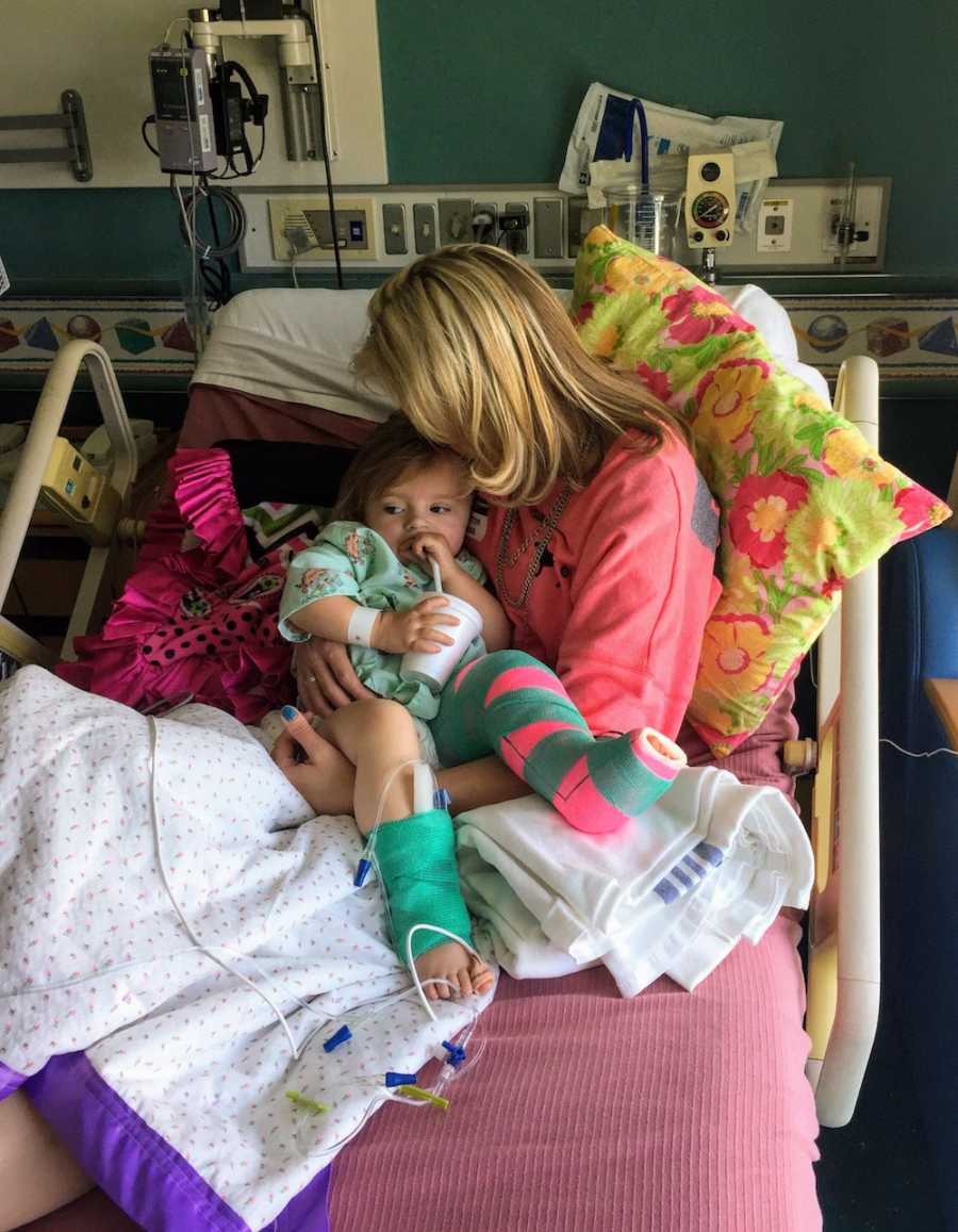 Mother sits in hospital bed holding daughter with cast on her leg in her arms