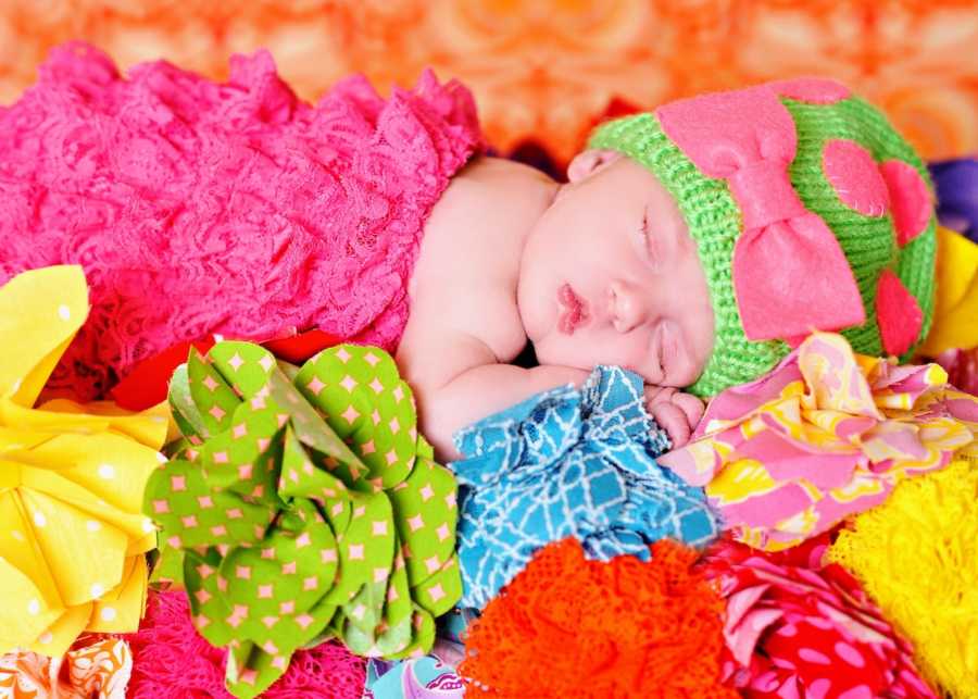 Baby girl in pink puffy dress and green hat lays asleep on colored and pattered paper flowers