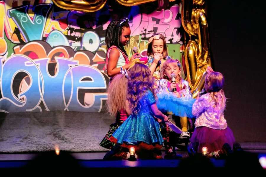 Little girl with Osteogenesis Imperfecta sits in wheelchair on stage singing with four girls at her side