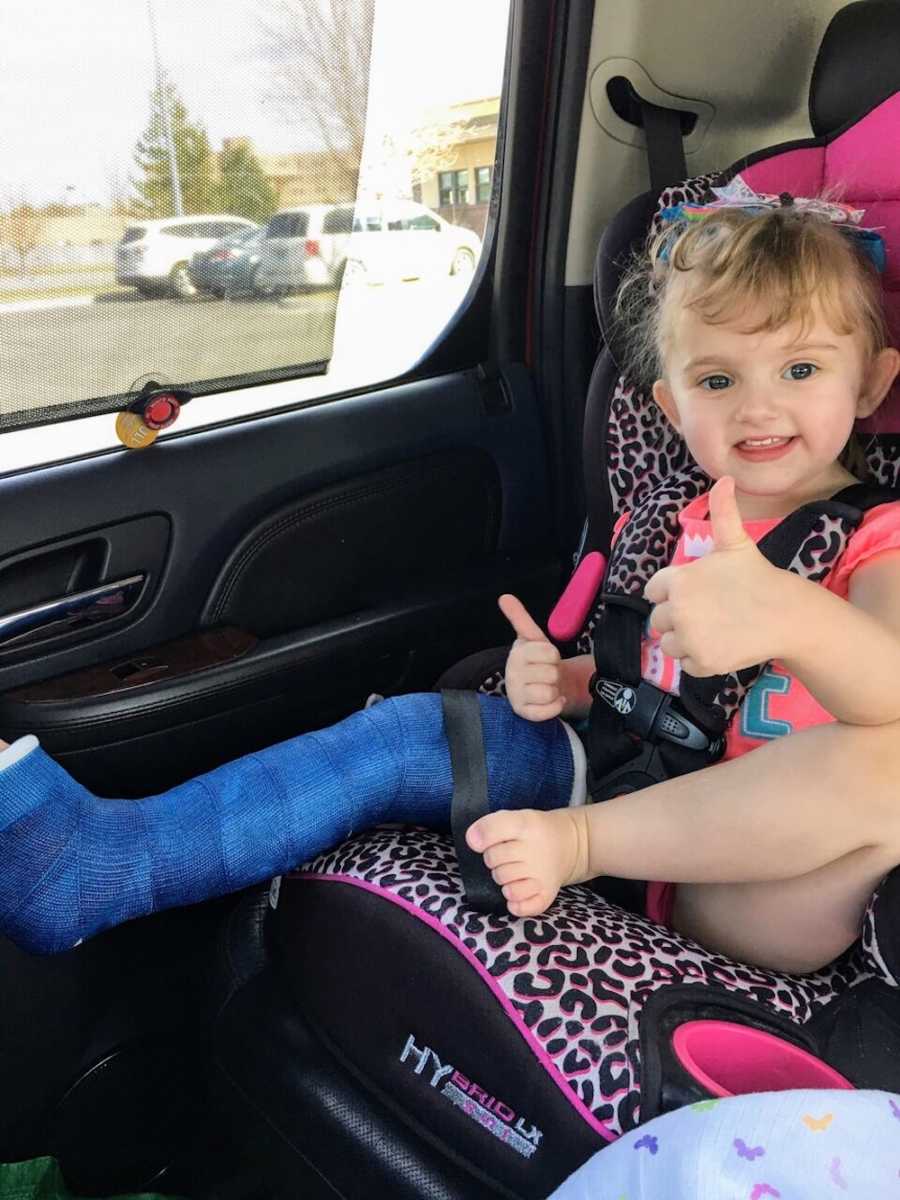 Little girl with Osteogenesis Imperfecta sits in carseat with two thumbs up and cast on her leg