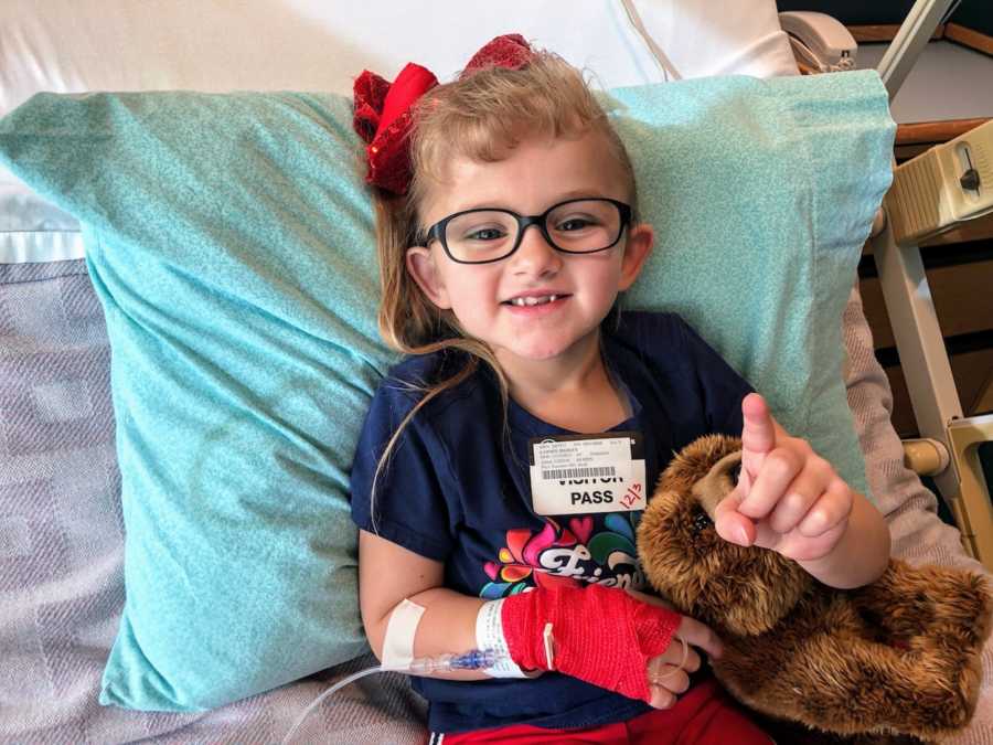 Little girl with Osteogenesis Imperfecta sits up in hospital bed smiling while holing up number one sign