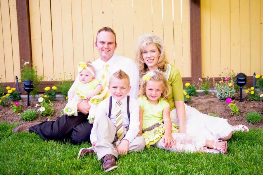 Husband and wife sit on ground outside in yard with their three children