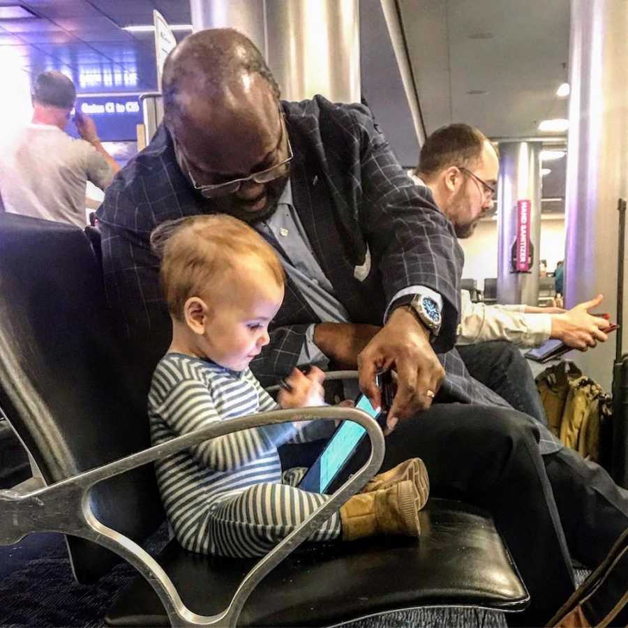 Little girl sits in chair in airport sitting beside man who helps her play on his tablet