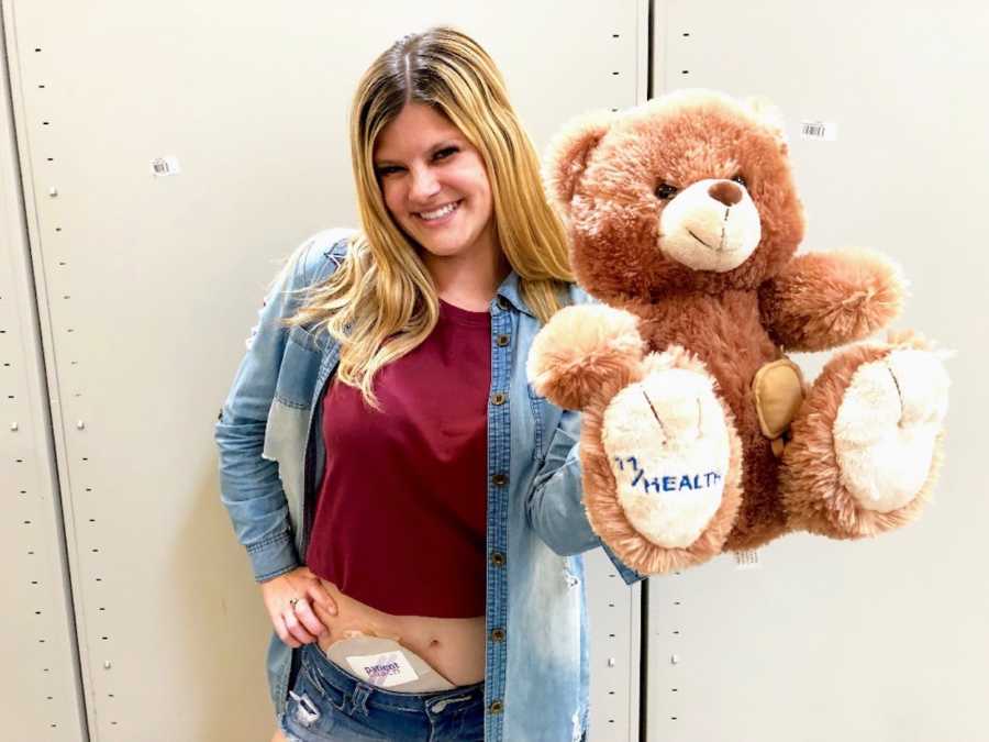 Woman stands on hand on hip with ostomy bag showing as she holds up teddy bear