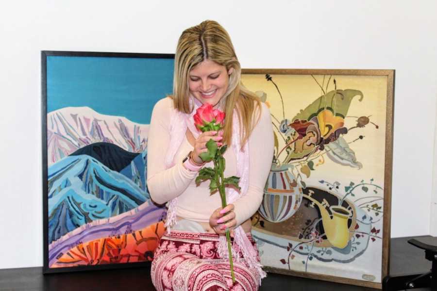 Woman with Crohn's disease and ostomy bag sits smiling in front of paintings as she looks down at flower in her hand