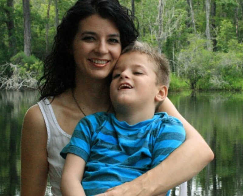 Mother smiles with son who has cerebral palsy on her lap with body of water in background 