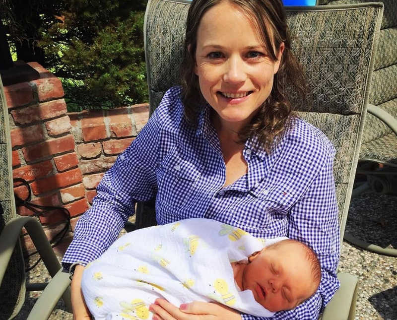 Mother sits outside in chair smiling as she holds sleeping baby in her lap