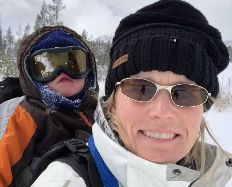 Mother wearing ski clothes smiles in selfie with young son swaddled to her back