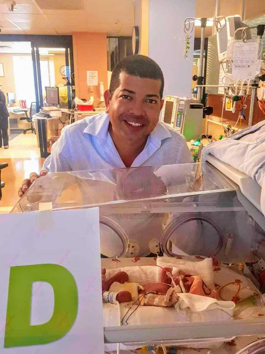Man smiles as he stands beside newborn in NICU