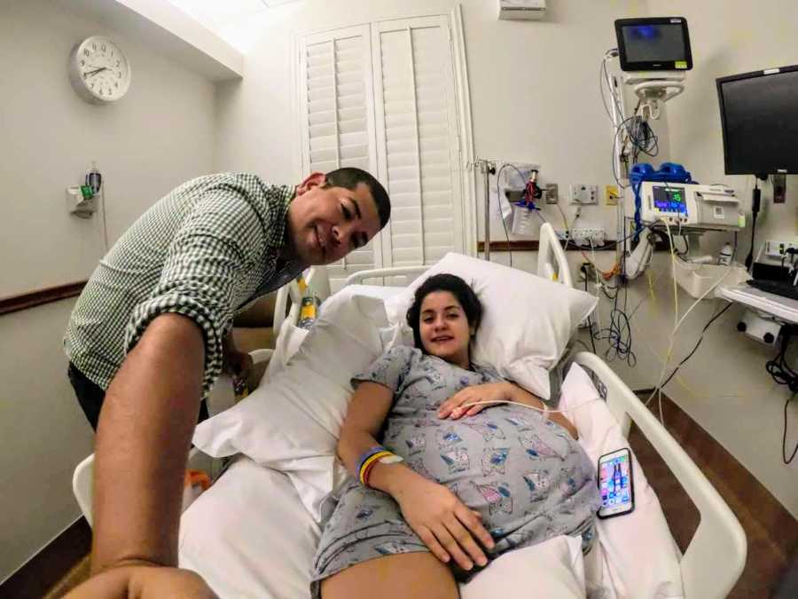 Woman pregnant with quadruplets lays in hospital bed as her husband stands smiling at her side