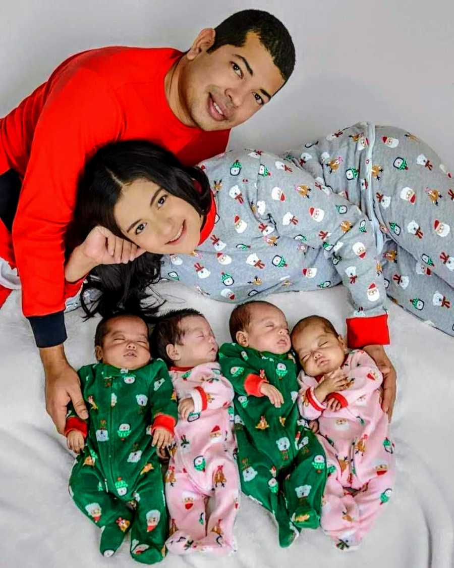 Husband and wife smiles as they lay on their sides next to quadruplet babies laying asleep in Christmas onesies