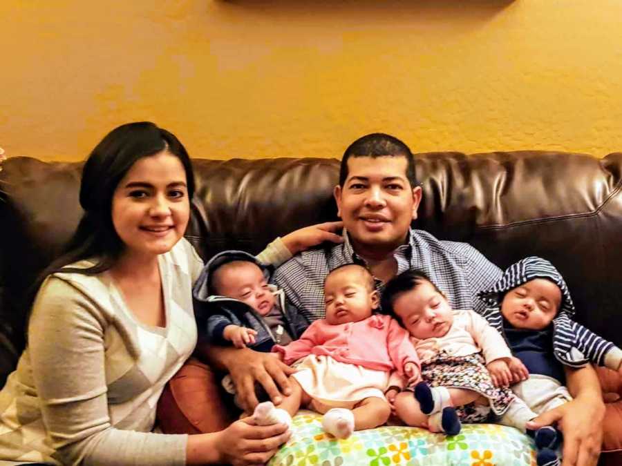 Mother smiles as she sits on couch beside husband who has quadruplet babies in his lap