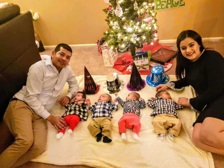 Husband and wife lay on floor beside baby quadruplets with new years hats by them
