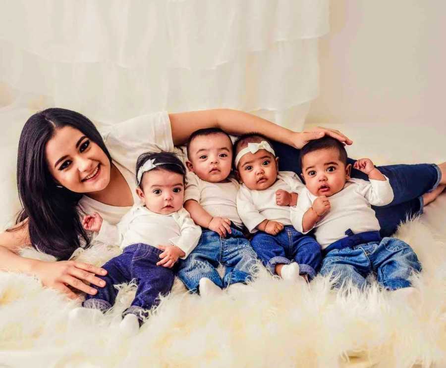 Mother smiles as she lays on her side with quadruplet babies sitting and leaning back on her