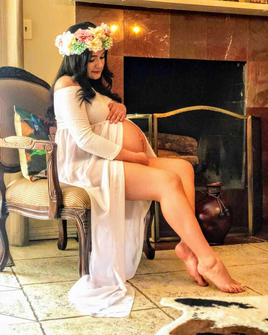 Pregnant woman sits in chair in home wearing flower crown as she holds her stomach