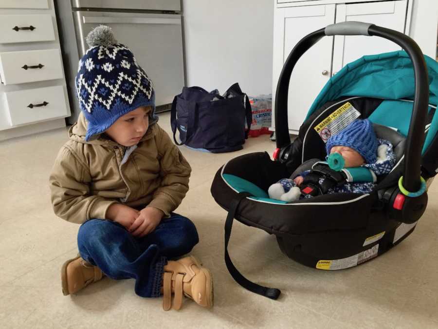 Little boy with autism sits on floor of kitchen looking at baby brother who lays asleep in car seat