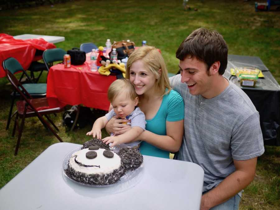 Father sits outside at table beside wife who has son in her lap in front of Mickey Mouse cake