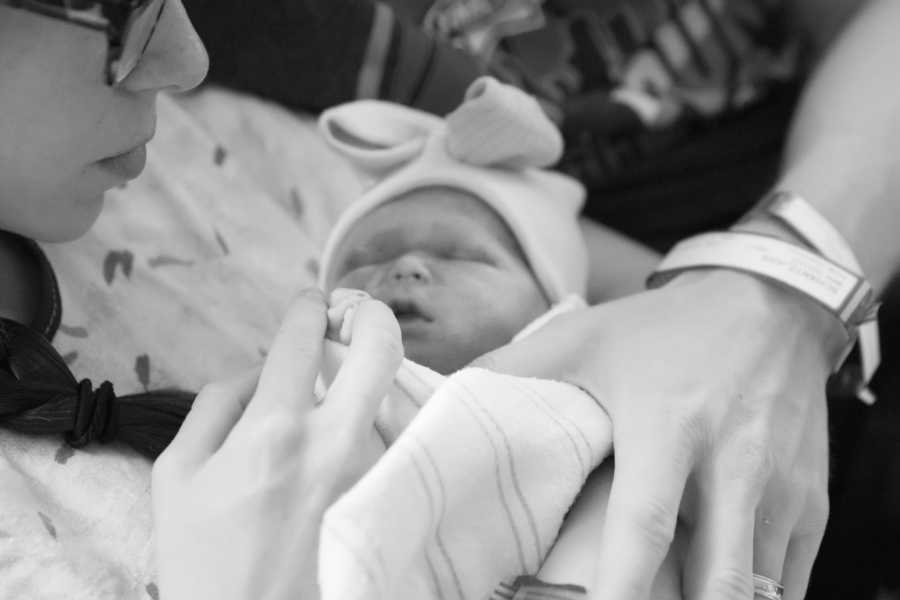 Newborn lays in mother arm holding onto her finger as father places his hand on her