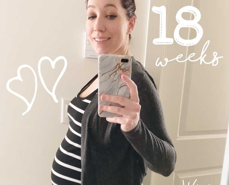 Pregnant woman stands smiling in mirror selfie with words "18 weeks" photoshopped on it