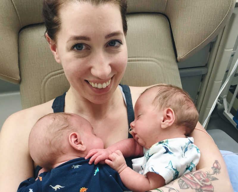Mother smiles in NICU with her twin baby boys asleep on her chest