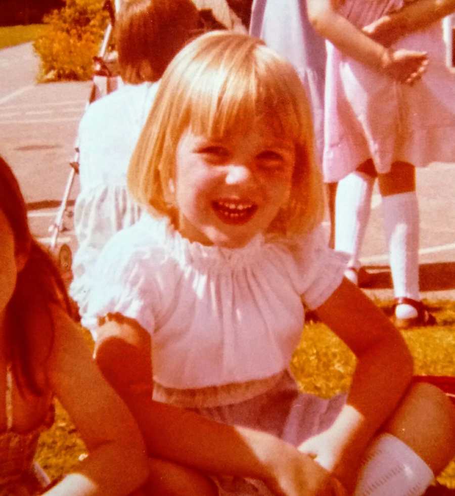 Little girl who will be abused later in life smiles as she sits on ground outside 