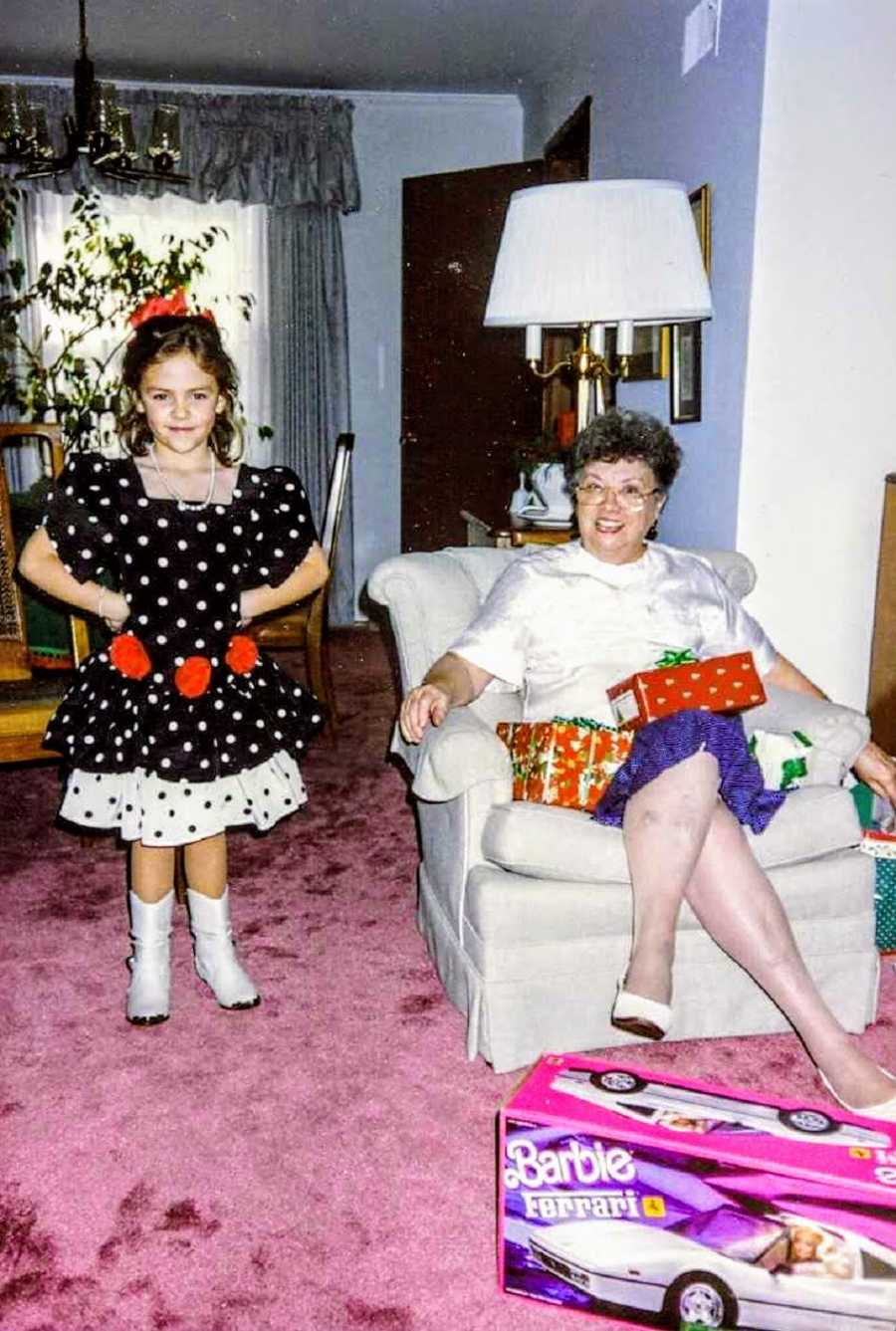 Little girl stands in dress in home beside grandmother she was named after who sits in chair beside her