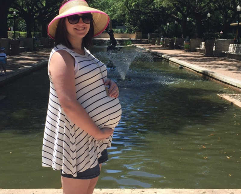 Pregnant woman stands beside fountain as she holds her stomach