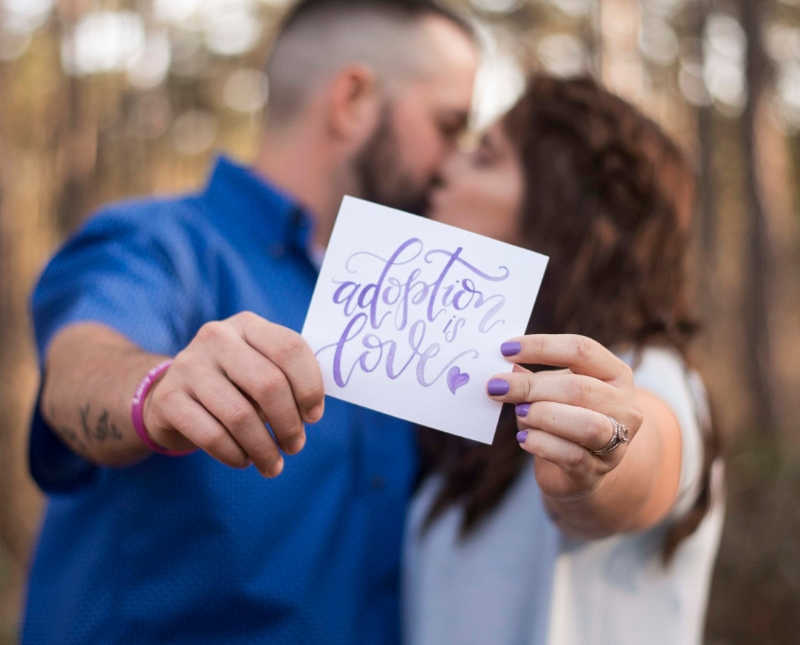 Husband and wife who lost their first born kiss while they hold paper that says, "adoption is love"