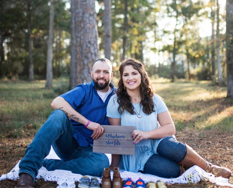 Husband and wife who lost their first born sit on blanket in wooded area holding wooden sign