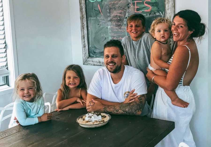 Woman who says her relationship with husband has changed after kids stands smiling beside table holding baby beside husband and three other kids