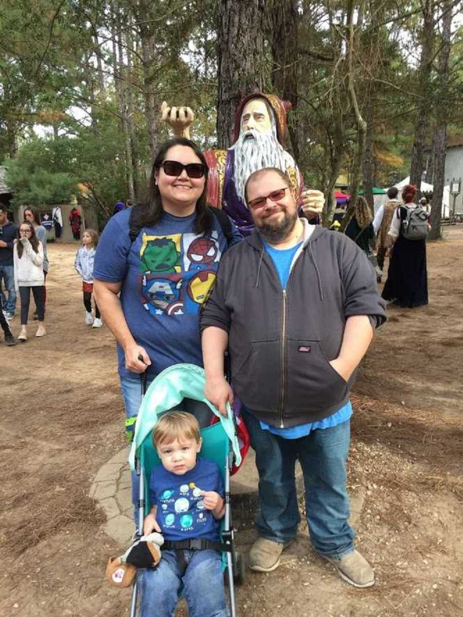 Husband and wife stand outside in wooded area with their toddler in stroller in front of wizard statue