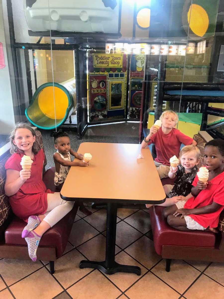 Five young siblings sit at table eating ice cream cone beside indoor play structure