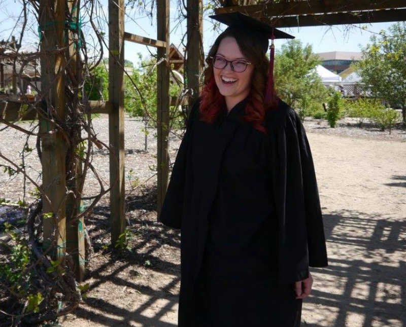 Young woman with Thoracic Outlet Syndrome stands outside in cap and gown smiling