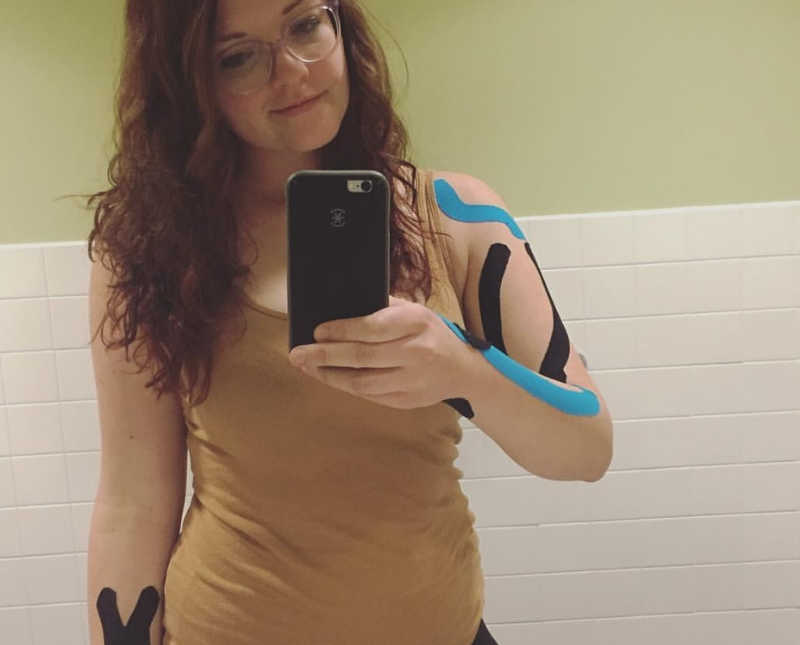 Woman with Thoracic Outlet Syndrome stands smiling in mirror selfie with KT tape on her arms